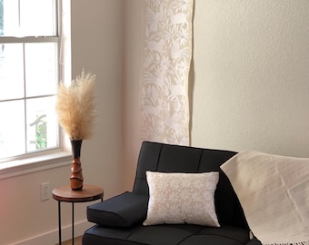 Op de afbeelding: Een zwarte futonbank met een beige kussen met bloemenpatroon en een witte plaid met franjes. Naast de bank staat een ronde houten bijzettafel met een bruine vaas met gedroogd pampasgras en een lange witte wandhanger met diermotieven.
