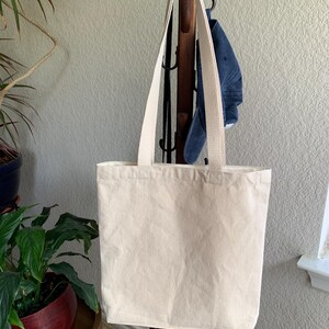 Op de afbeelding: Een cr&egrave;mekleurige draagtas met bijpassende bandjes hangt aan een houten kapstok. De tas is gemaakt van stevig canvas materiaal. Een blauwe denim pet hangt achter de tas. Een plant in een pot is zichtbaar op de voorgrond.