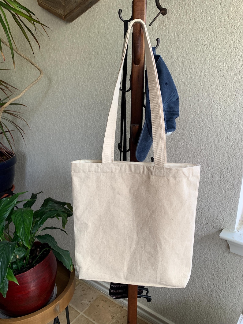 Op de afbeelding: Een cr&egrave;mekleurige draagtas met bijpassende bandjes hangt aan een houten kapstok. De tas is gemaakt van stevig canvas materiaal. Een blauwe denim pet hangt achter de tas. Een plant in een pot is zichtbaar op de voorgrond.
