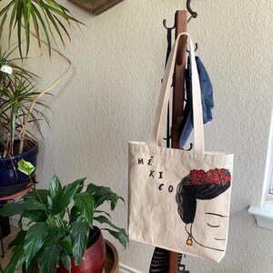 Op de afbeelding: Beige draagtas met een Frida Kahlo-achtige illustratie, bloemenkrans en het woord "M&Eacute;XICO". De tas heeft cr&egrave;mekleurige bandjes en hangt aan een houten kapstok. Een spijkerbroek hangt er ook aan.