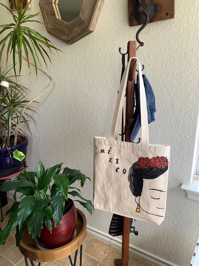 Op de afbeelding: Beige draagtas met een Frida Kahlo-achtige illustratie, bloemenkrans en het woord "M&Eacute;XICO". De tas heeft cr&egrave;mekleurige bandjes en hangt aan een houten kapstok. Een spijkerbroek hangt er ook aan.