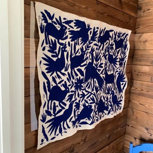 Puede incluir: Textil rectangular blanco roto con un diseño bordado en azul oscuro de pájaros, conejos y follaje. El textil está montado en una pared de madera, mostrando un estilo de arte popular.