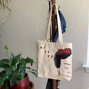 Op de afbeelding: Beige draagtas met een gestileerde illustratie van een vrouw met een bloemenkrans en het woord "M&Eacute;XICO". De tas heeft cr&egrave;mekleurige handvatten en hangt aan een houten kapstok. Een potplant staat op de voorgrond.