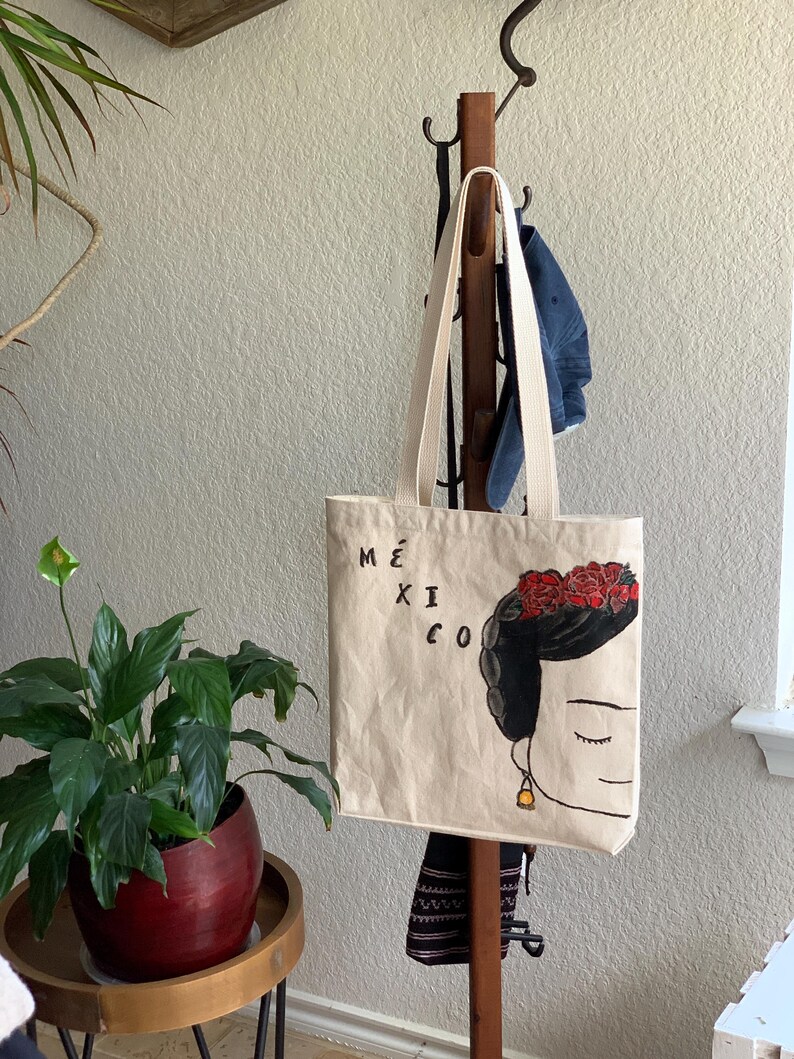Op de afbeelding: Beige draagtas met een gestileerde illustratie van een vrouw met een bloemenkrans en het woord "M&Eacute;XICO". De tas heeft cr&egrave;mekleurige handvatten en hangt aan een houten kapstok. Een potplant staat op de voorgrond.