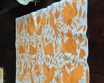 Op de afbeelding: Een rechthoekige, lichtblauwe tafelloper met een geborduurd oranje ontwerp met vogels, bloemen en botanische elementen. De loper ligt op een donkerbruine houten tafel. Een boeket kleurrijke bloemen staat op de achtergrond.
