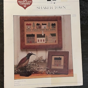 May include: Cross stitch pattern for a Shaker Town scene with houses, trees, and the text "May peace bless our home". The pattern is from The Need'l Love Company.