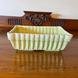May include: A pale yellow ceramic planter with a fluted design. The planter has a rectangular shape and is sitting on a wooden surface.