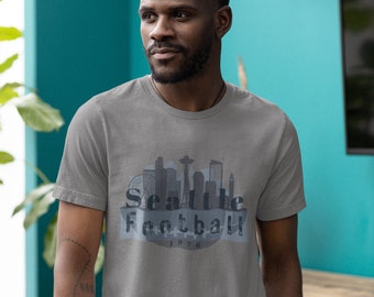 以下が含まれることがあります： シアトルのスカイラインのデザインと「Seattle Football 1976」の文字が入ったグレーのTシャツ。デザインは青とグレーの色合いです。Tシャツはクルーネックです。