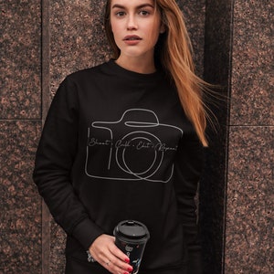 Könnte beinhalten: Schwarzes Sweatshirt mit einem weißen Umriss einer Kamera und dem Text "Shoot.  Edit. Repeat." Das Model hält eine schwarze Kaffeetasse mit einem Blumenmuster.