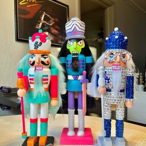 May include: Three decorative nutcrackers standing on rectangular bases. The left one features teal hair, a white uniform with teal accents, and a pink staff. The center one has green skin, a purple hat, and a pink sword. The right one is covered in blue sequins.