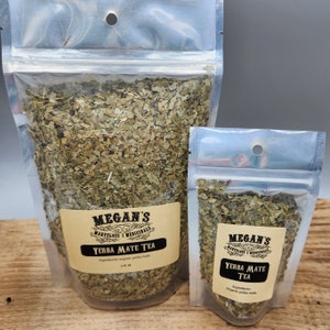 Two clear plastic bags filled with loose green yerba mate tea leaves. The larger bag has a label that reads "Megan's Marvelous Medicinals Yerba Mate Tea" and the smaller bag has a label that reads "Megan's Marvelous Medicinals Yerba Mate Tea".