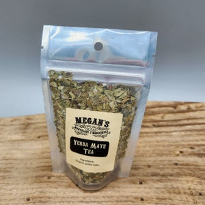 A clear plastic bag filled with loose green yerba mate tea leaves. The bag has a label that reads "Megan's Marvelous Medicinals Yerba Mate Tea" and "Ingredients: Organic yerba mate."