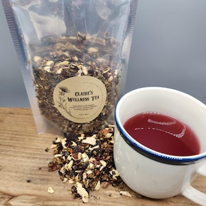 May include: A clear bag of Claire's Wellness Tea with a round label, next to a white mug filled with red tea. Loose tea leaves are scattered on a wooden surface. The label lists ingredients: juniper berry, hibiscus flowers, rosehips, and ginger root.