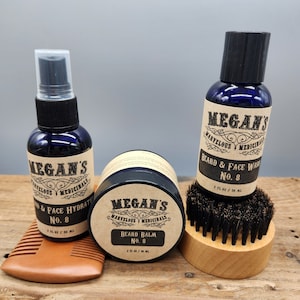 May include: A set of beard care products in blue bottles and a jar with black lids. The products are labeled "Megan's Marvelous Medicinals" and include a beard balm, beard and face wash, and beard and face hydrator. The bottles are 2 fl oz / 59 ml. The products are arranged on a wooden surface with a wooden brush.
