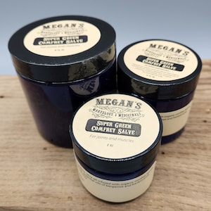 May include: Three jars of Super Green Comfrey Salve, a natural remedy for joints and muscles. The jars are labeled with the product name and instructions for use. The jars are on a wooden surface.