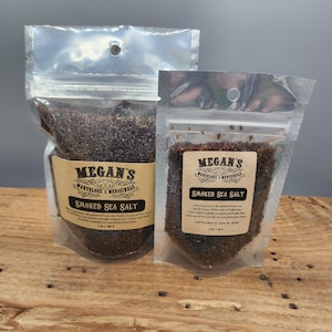May include: Two clear plastic pouches filled with dark brown smoked sea salt. Each pouch has a label with the text "Megan's Marvelous Medicinals Smoked Sea Salt". The pouches are on a wooden surface.