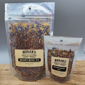 May include: Two clear plastic bags filled with loose leaf tea. The larger bag is labeled "Megan's Marvelous Medicinals Dragon's Hoard Tea" and lists the ingredients as rooibos, rosehips, elderflowers, calendula, and cornflower. The smaller bag is labeled "Megan's Marvelous Medicinals Dragon's Hoard Tea" and lists the ingredients as rooibos, rosehips, elderflowers, calendula, and cornflower.