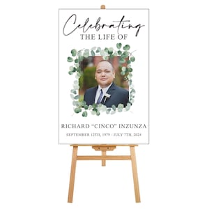May include: A white sign with a black border and a green eucalyptus wreath surrounding a photo of a man. The text reads "Celebrating the Life of Richard "Cinco" Inzunza September 12th, 1979 - July 7th, 2024".