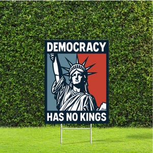 May include: A rectangular yard sign featuring the Statue of Liberty with the words "DEMOCRACY" above and "HAS NO KINGS" below. The background is split into blue and red sections. The sign is mounted on a metal stake and placed on a grassy lawn.