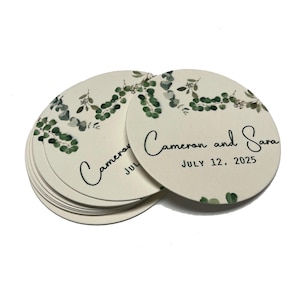 May include: A stack of round, white paper coasters with a green watercolor eucalyptus design. The top coaster has the text "Cameron and Sara" and "JULY 12, 2025" printed in black.