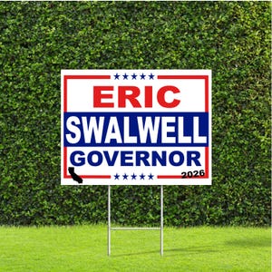 May include: A white and red campaign sign with the text "ERIC SWALWELL GOVERNOR 2026" in red and blue. The sign is placed in a grassy yard with a green hedge background.