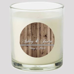 May include: A clear glass candle jar with a brown woodgrain label. The label has the text "Ashley & Justin" and "May 5, 2018" in white.
