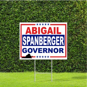 May include: A white and red sign with the text "ABIGAIL SPANBERGER GOVERNOR" in blue and red lettering. The sign includes a silhouette of the state of Virginia and is mounted on a metal stand, placed on a grassy lawn.