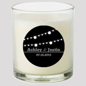 May include: A white candle in a clear glass jar with a black and white circular label. The label has a design of white string lights and the text "Ashley & Justin 07.18.2015".