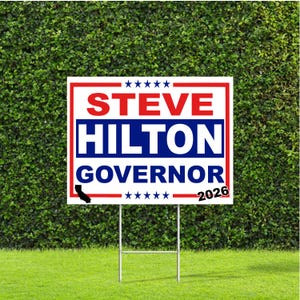 May include: A white and red campaign sign with the text "STEVE HILTON GOVERNOR 2026" in bold blue and red lettering. The sign includes a California outline and star graphics, mounted on a metal frame, set against a green lawn and hedge.