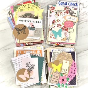 May include: A collection of scrapbook ephemera, including paper cutouts, tags, and decorative elements. The assortment features butterfly and floral motifs, with text that reads "POSITIVE VIBES" and "Guest Check". Various colors are present, including yellow, pink, and teal.