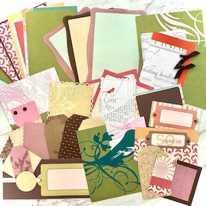 May include: A collection of scrapbook paper and embellishments in various shapes and sizes. Includes tags, cards, and patterned papers in shades of green, pink, brown, and cream. Some pieces feature decorative edges, while others have printed text and illustrations.