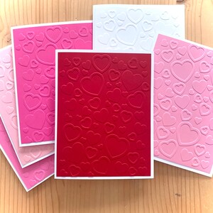 May include: A collection of handmade greeting cards with embossed heart patterns. The cards feature various shades of pink, red, and white, with a white border. The hearts are of different sizes, creating a textured effect. The cards are arranged on a wooden surface.