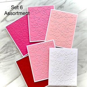 May include: A collection of embossed greeting cards in various shades of pink and red, featuring a heart pattern. The cards are arranged on a light-colored surface. The text "Set 6 Assortment" is visible in the upper left corner.