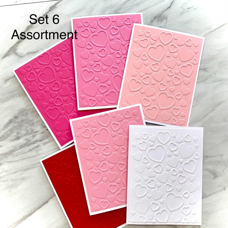 May include: A collection of embossed greeting cards in various shades of pink and red, featuring a heart pattern. The cards are arranged on a light-colored surface. The text "Set 6 Assortment" is visible in the upper left corner.