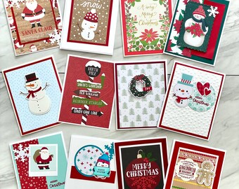 May include: A collection of Christmas greeting cards with various designs. Santa Claus, snowmen, and festive text are featured. The cards have red, white, and green accents, with some cards featuring snowflakes and holiday imagery.