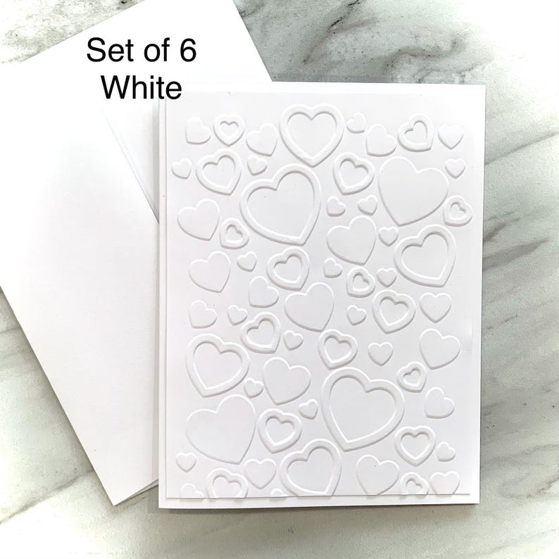 May include: Set of six white greeting cards with embossed heart designs. Each card features a pattern of various sized hearts. Includes white envelopes. Text on the image reads "Set of 6 White".