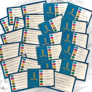 May include: A collection of blue and gold trivia cards with the word "Genus" in gold script. Each card features questions and answers, with colored letters indicating the answer's category. The cards are scattered on a white surface.