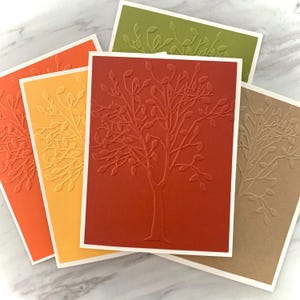 May include: A collection of handmade greeting cards in various fall colors. Each card features an embossed tree design with a white border. Colors include orange, yellow, red, green, and brown. The cards are arranged in a fan shape, showcasing the different colors and designs.