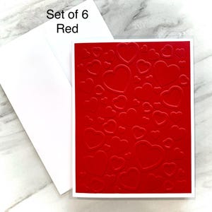 May include: A set of six red greeting cards with a white border and matching envelopes. The cards feature a textured pattern of embossed hearts. The text "Set of 6 Red" is in the upper left corner.