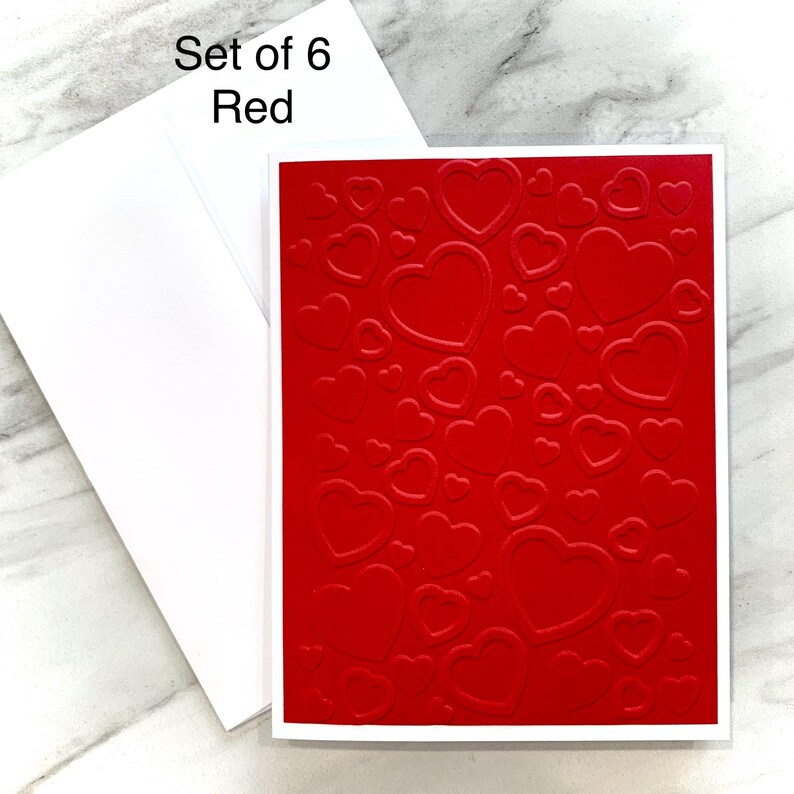 May include: A set of six red greeting cards with a white border and matching envelopes. The cards feature a textured pattern of embossed hearts. The text "Set of 6 Red" is in the upper left corner.