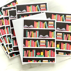 May include: Greeting cards with a bookshelf design. The cards feature a black background with colorful books labeled with subjects like Algebra, History, and Science. White envelopes are included.