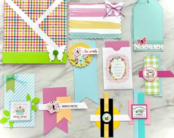 May include: A collection of colorful paper crafts, including tags, bookmarks, and decorative elements. Designs feature butterflies, floral motifs, and text like "Moments Matter" and "Live Simply." The color palette includes pink, green, blue, and yellow.