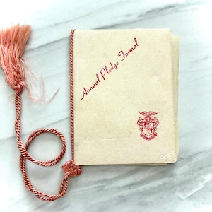 May include: A vintage booklet with the words "Annual Pledge Formal" in red script. The booklet has a red and pink tassel attached to a pink cord. A red crest is printed on the booklet.