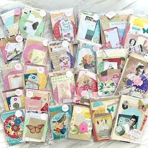 May include: A collection of assorted paper ephemera packs, each sealed in a clear bag. The packs contain decorative tags, cards, and paper elements with floral, butterfly, and vintage-inspired designs. Some packs have price tags.