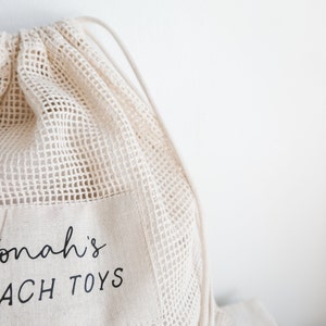 May include: A white mesh drawstring bag with a white fabric pocket that says "Jonah's Beach Toys" in black text.