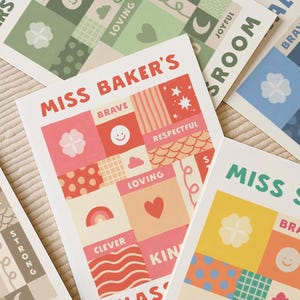 May include: A collection of rectangular art prints featuring a grid of squares with various patterns and words including "MISS BAKER'S", "BRAVE", "RESPECTFUL", "LOVING", "CLEVER", and "KIN". The designs incorporate icons like hearts, rainbows, stars, and smiley faces in red, pink, orange, yellow, green, and blue color palettes.