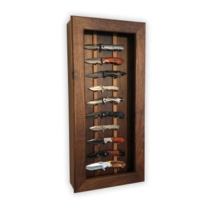 May include: A wooden wall-mounted display case with a dark brown finish, featuring a collection of eight pocket knives displayed on a light brown background.