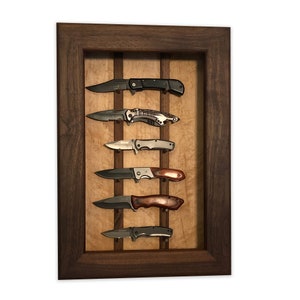 May include: A wooden frame with a light wood background and five pocket knives displayed on the wall. The knives are arranged vertically and are held in place by small metal hooks. The frame is made of dark wood and has a simple design.