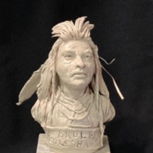 Könnte beinhalten: Eine weiße Keramikbüste eines indianischen Mannes mit langen Haaren und Federn. Die Büste steht auf einem weißen Marmorsockel mit der Inschrift "BRULE LITTLE HAWK".