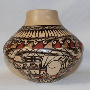 Può includere: Un vaso in ceramica color beige con disegni geometrici neri e rossi. Il vaso ha un corpo arrotondato e un collo stretto.